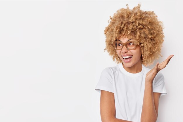 Plan horizontal d'une femme bouclée folle réagit émotionnellement sur quelque chose qui garde les sourires levés de la paume porte joyeusement un t-shirt décontracté et des lunettes pose sur fond blanc copiez l'espace pour votre promo