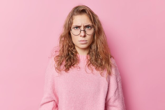 Plan horizontal d'une femme européenne déçue en colère regarde malheureusement la caméra a une expression grincheuse entend quelque chose de désagréable porte des lunettes rondes et un cavalier isolé sur fond rose
