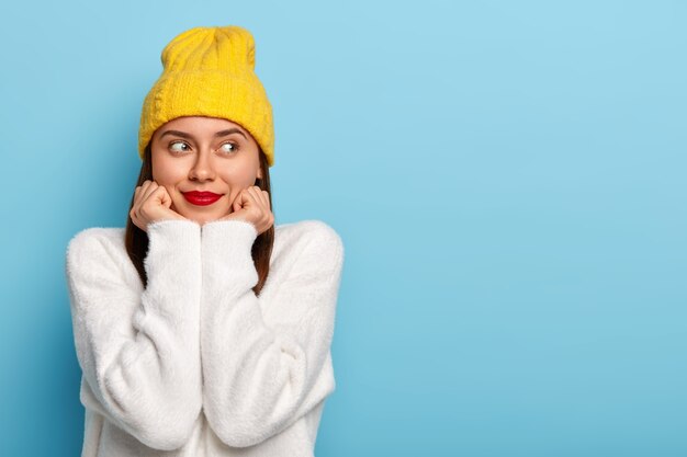 Plan horizontal d'une femme européenne rêveuse heureuse porte un maquillage minimal, rouge à lèvres, regarde de côté, vêtu d'un chapeau jaune et d'un pull blanc, pose sur fond bleu, fasciné et heureux