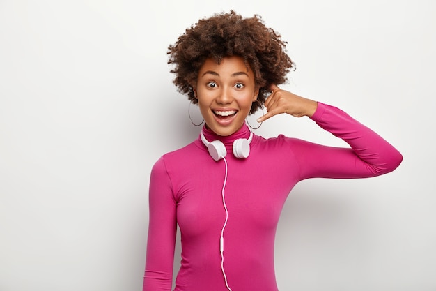 Plan horizontal d'une femme heureuse à la peau sombre fait un geste d'appel, regarde positivement, porte des écouteurs autour du cou, pose sur fond blanc, se sent heureux. Le langage du corps