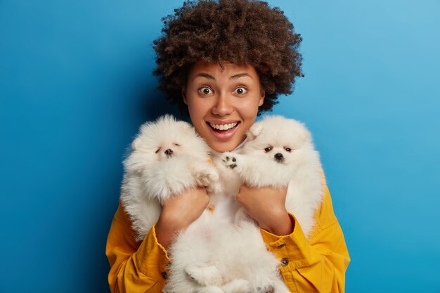 Plan horizontal d'une femme joyeuse bouclée tient fermement deux chiots de race avec fourrure blanche, bénéficie d'un cadeau de petit ami