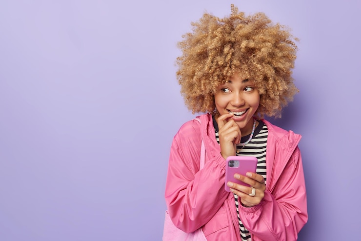 Photo gratuit plan horizontal d'une femme joyeuse et rêveuse regarde ailleurs vêtue d'une veste rose porte un sac utilise un téléphone portable pour discuter et surfer sur internet isolé sur fond violet espace vide pour le texte