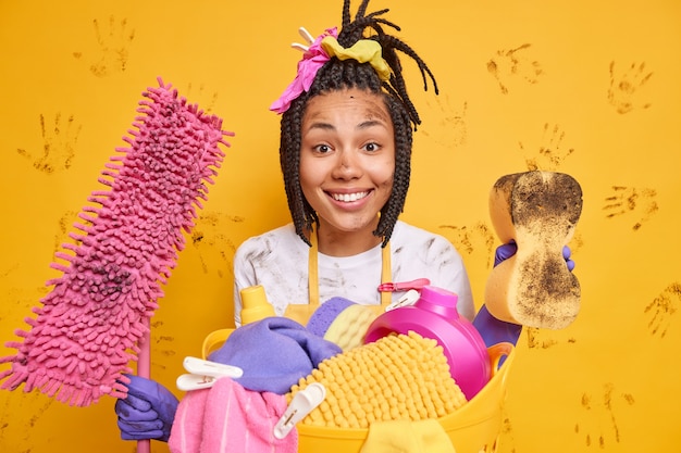 Plan horizontal d'une femme de ménage heureuse souriant agréablement de bonne humeur après avoir fini de nettoyer l'appartement pose près d'un panier à linge isolé sur un mur jaune