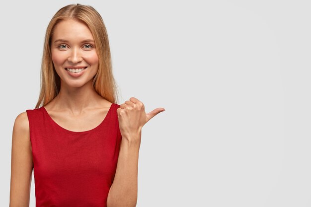 Plan horizontal d'une femme mince assez heureuse a un sourire doux, un regard attrayant, des points avec le pouce de côté, vêtu d'un t-shirt rouge