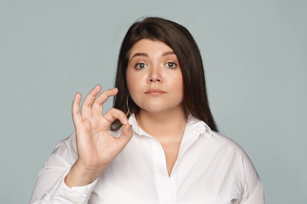 Plan horizontal d'une femme potelée en surpoids grave et confiante, vêtue d'une chemise formelle blanche reliant l'avant et le pouce, faisant un geste correct, montrant que tout est sous contrôle