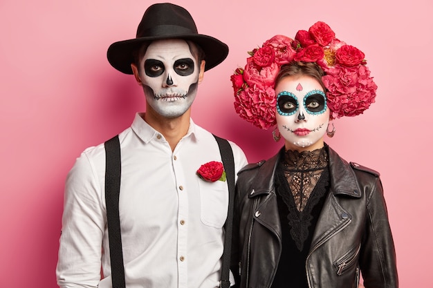 Plan horizontal d'une femme sérieuse et d'un homme vêtu de costumes d'Halloween, porter du maquillage squelette, guirlande de pivoines