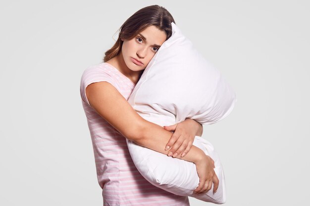 Plan horizontal d'une femme triste sérieuse porte un oreiller blanc de près, porte un pyjama, regarde directement la caméra, pose sur le blanc, veut se reposer, prêt à dormir, fait de beaux rêves