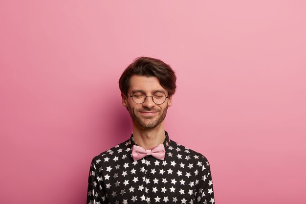 Plan horizontal d'un gars hipster heureux se tient les yeux fermés, pense à quelque chose de positif, a un sourire sur le visage, des soies sombres