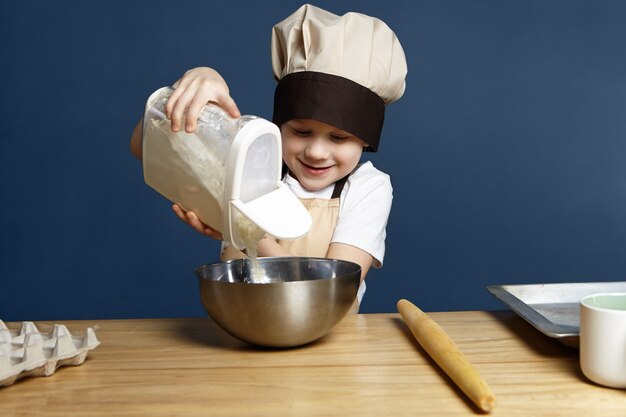 Plan horizontal de l'heureux enfant européen de 8 ans ayant l'air excité alors qu'il verse de la farine de blé dans un bol en métal tout en allant faire de la pâtisserie par lui-même, debout au mur de la cuisine vide