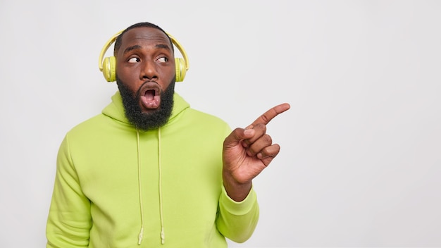 Plan horizontal d'un homme barbu choqué à la peau foncée écoute une piste audio via un casque sans fil porte un sweat-shirt vert sur un espace vide isolé sur un mur blanc