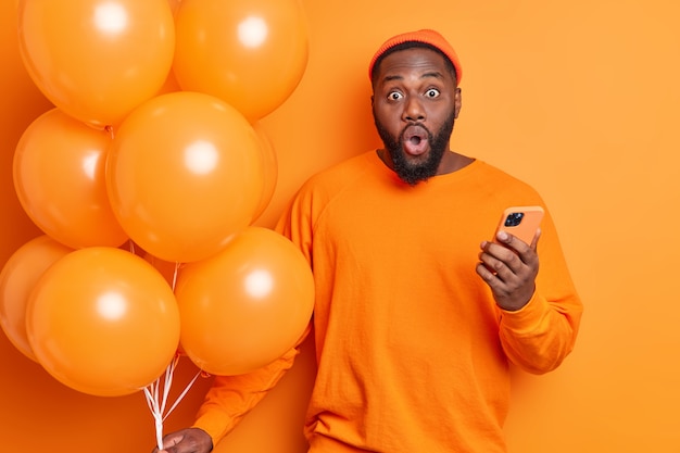 Plan horizontal d'un homme barbu choqué regarde de façon surprenante la caméra habillée avec désinvolture tient un téléphone portable et un tas de ballons en partie isolée sur un mur orange