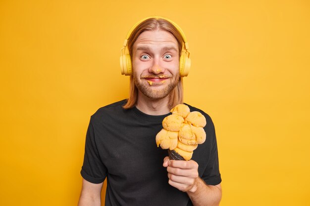 Plan horizontal d'un homme barbu positif avec de longs cheveux roux, le visage barbouillé de crème glacée tient une grosse glace jaune vêtue avec désinvolture s'amuse profite des vacances d'été écoute de la musique avec des écouteurs