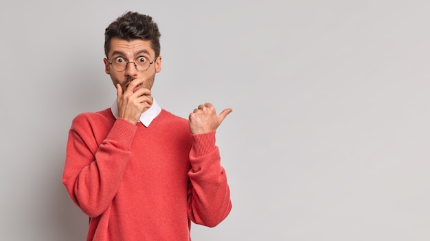 Plan horizontal d'un homme choqué couvre la bouche regarde la caméra avec des yeux buggés pointe le pouce sur un espace vide