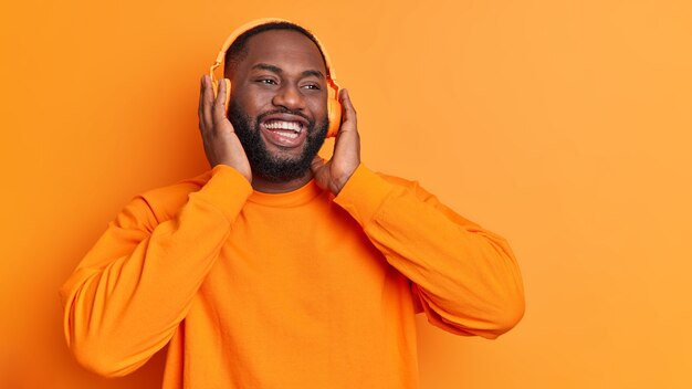 Plan horizontal de l'homme heureux garde la main sur un casque stéréo sourit largement apprécie la chanson agréable passe du temps libre à écouter de la musique isolée sur un mur orange vif avec un espace vide