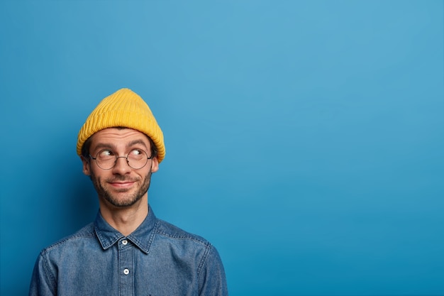 Plan horizontal d'un homme heureux et joyeux regarde loin avec une expression de rêve, a l'intention de faire quelque chose, vêtu de vêtements élégants