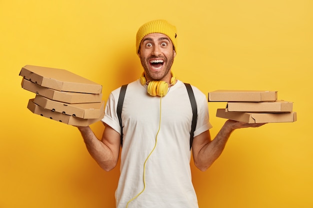 Plan Horizontal De L'homme Heureux Tient Deux Piles De Boîtes En Carton Avec Pizza, A Surpris L'expression Joyeuse, Travaille Comme Courrier Dans Un Restaurant Local