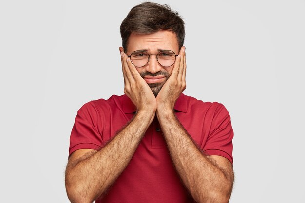 Plan horizontal d'un homme mécontent aux cheveux noirs, touche les joues avec les mains, a une expression faciale négative, porte un t-shirt décontracté rouge, se tient contre un mur blanc. Concept de personnes et d'émotions