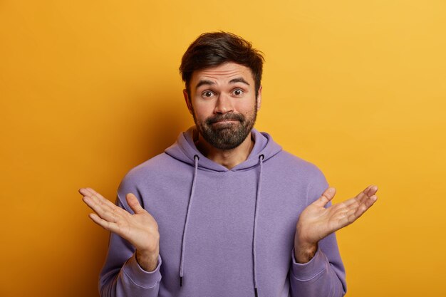 Plan horizontal d'un homme de race blanche barbu inconscient étend les paumes sur le côté, hausse les épaules, fait face au dilemme, fait un choix, porte un sweat-shirt décontracté, regarde avec le doute, pose contre le mur jaune