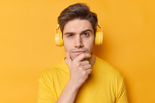 Plan horizontal d'un homme réfléchi tient le menton et regarde sérieusement la caméra écoute sa chanson préférée via un casque stéréo vêtu de vêtements décontractés pose sur fond jaune vif Laissez-moi réfléchir