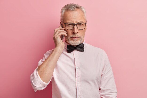 Plan horizontal d'un homme senior parle au téléphone avec une expression sérieuse, tient un téléphone mobile moderne près de l'oreille, a une conversation