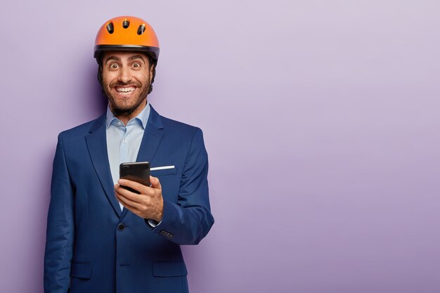 Plan horizontal d'un ingénieur professionnel heureux porte un costume formel et un casque de protection orange, utilise un smartphone pour contrôler le travail en ligne sur le chantier de construction