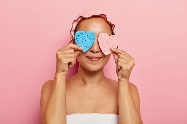 Plan horizontal intérieur d'une jeune femme asiatique ravie couvre les yeux avec deux éponges en forme de cœur, s'amuse après la douche, utilise des produits cosmétiques pour appliquer la crème, a une peau lisse et foncée.