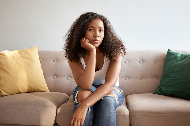 Plan horizontal d'une jeune femme africaine à la mode avec une coiffure volumineuse ayant un look ennuyé, doit rester à la maison à cause du mauvais temps pluvieux, assise seule sur un canapé gris. Les gens et le style de vie