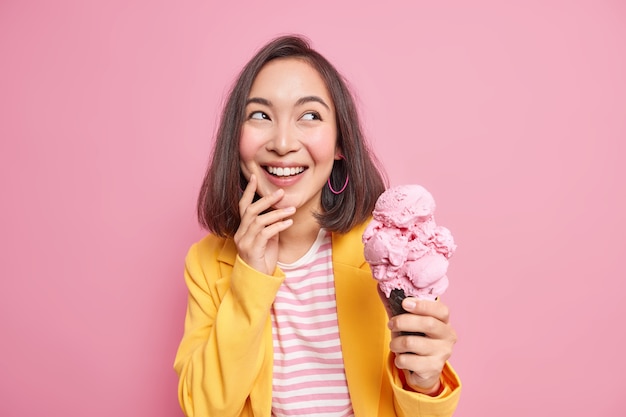 Plan horizontal d'une jeune femme asiatique brune et joyeuse qui détourne le regard avec plaisir tient une délicieuse crème glacée froide dans une gaufre porte des vêtements élégants et profite des vacances d'été isolées sur un mur rose.