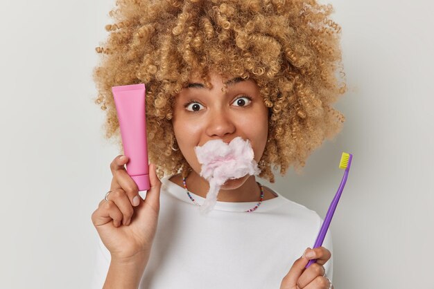 Plan horizontal d'une jeune femme aux cheveux bouclés a la bouche pleine de barbe à papa tient un tube de dentifrice et une brosse à dents prend soin des dents vêtues d'un t-shirt décontracté isolé sur fond blanc