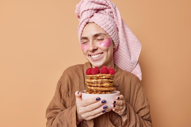 Plan horizontal d'une jeune femme heureuse ferme les yeux du plaisir tient de délicieuses crêpes au sirop d'érable sourit agréablement vêtue d'un costume de sommeil subit des procédures de beauté après avoir pris une douche