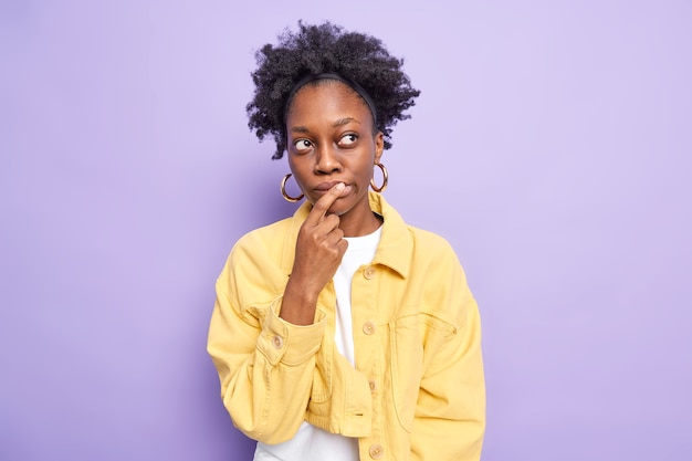 Plan horizontal d'une jeune femme pensive à la peau foncée fait le choix, garde l'index sur les lèvres et réfléchit profondément à quelque chose