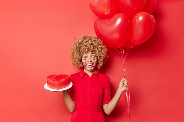 Plan horizontal d'une jeune femme positive surprise reçoit le cadeau de son petit ami le jour de la Saint-Valentin tient un gâteau en forme de coeur et des ballons porte une robe profite de vacances préférées isolées sur un mur de studio rouge