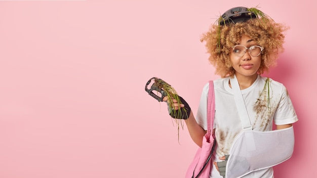 Photo gratuite plan horizontal d'une jeune femme réfléchie aux cheveux bouclés s'est cassé le bras après une conduite dangereuse à vélo s'arrête sur la route tient la pédale a panne de transport porte un équipement de protection pose à l'intérieur