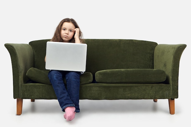 Plan horizontal d'une jeune fille européenne sérieuse de 10 ans, assise sur un canapé dans le salon, se gratter le visage, regardant la caméra avec une expression confuse pensive.
