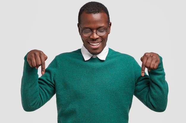 Plan horizontal d'un jeune homme afro-américain positif pointe les deux index vers le bas, étant assuré de son choix, a un sourire à pleines dents