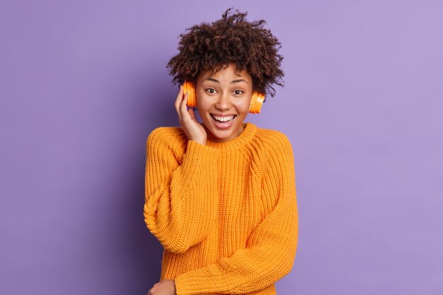 Plan horizontal d'une jolie femme afro-américaine aux cheveux bouclés glousse positivement écoute une mélodie agréable porte un casque stéréo porte un pull orange tricoté