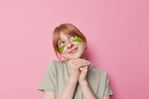 Plan horizontal d'une jolie femme rêveuse garde les mains sous le menton regarde au-dessus avec une expression satisfaite porte un t-shirt décontracté rêve de quelque chose tout en subissant des procédures de beauté pose à l'intérieur