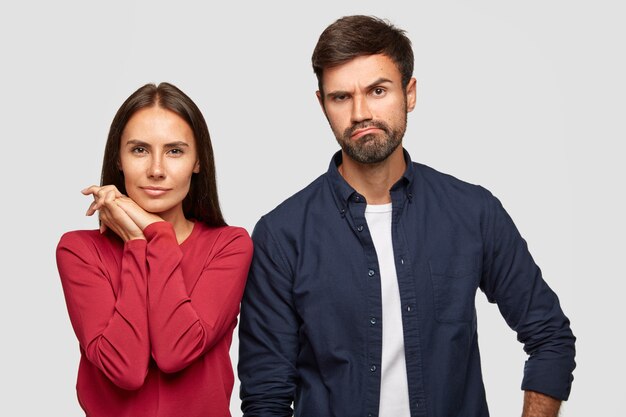 Plan horizontal de la jolie jeune femme et de son petit ami regardent sérieusement et de manière déroutante les prix en magasin, faire du shopping ensemble, vêtus de vêtements décontractés, isolés sur un mur blanc.