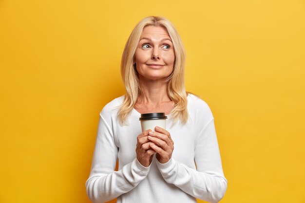 Plan horizontal d'une mamie réfléchie rêveuse a une pause-café profite du temps libre se souvient d'agréables souvenirs de sa jeunesse pose avec une tasse de boisson chaude habillée avec désinvolture