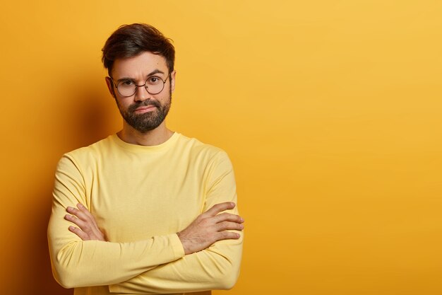 Plan horizontal d'un modèle masculin à l'air sérieux garde les bras croisés, n'aime pas faire du shopping, se sent ennuyé de passer du temps avec sa femme qui achète, porte des lunettes et un pull, isolé sur un mur jaune