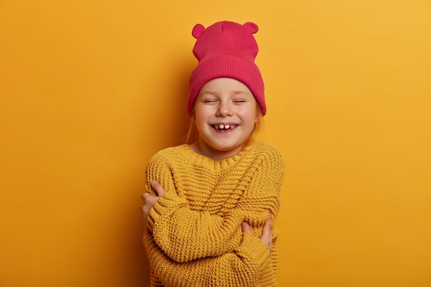 Plan horizontal d'une petite fille heureuse s'embrasse, se sent bien, porte un chapeau rose et un pull en tricot, étant de bonne humeur, isolée sur un mur jaune. Enfants, estime de soi