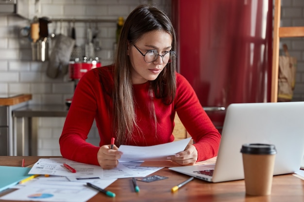 Plan horizontal d'un travailleur indépendant occupé lit des données d'information sur des papiers, vérifie les chiffres sur un ordinateur portable, étudie des graphiques sur des documents, s'assoit au bureau dans un intérieur de cuisine confortable.