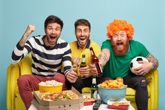 Plan horizontal de trois hommes joyeux se réunissent le week-end pour regarder un match de football, célèbre but de score, s'asseoir sur un canapé jaune, isolé sur un mur bleu. Gens, concept d'excitation