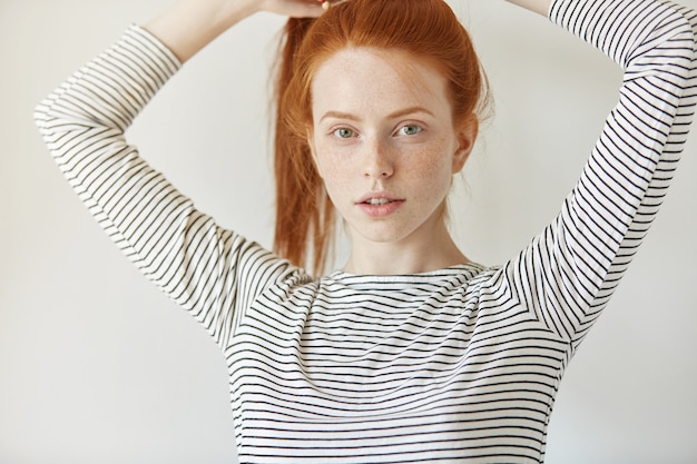 Plan Intérieur D'une Belle Adolescente Aux Taches De Rousseur Aux Longs Cheveux Roux Rassemblés En Queue De Cheval, Habillée En Passant, Posant à L'intérieur, Ajustant Sa Coiffure Avant D'aller à L'université. Concept De Jeunesse Et De Tendresse