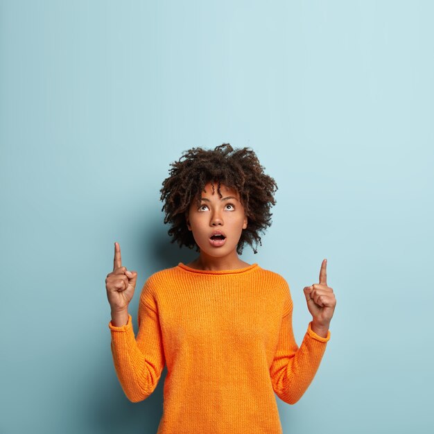 Plan intérieur d'une belle femme noire étonnée et surprise qui pointe vers le haut, montre la direction au-dessus, porte un pull orange, isolé sur un mur bleu. Objet de promots féminin à la peau sombre impressionnée