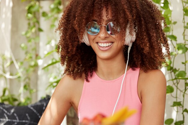 Plan intérieur d'une belle jeune femme avec une coiffure afro, porte des lunettes de soleil à la mode, écoute ses chansons préférées dans des écouteurs seul, a un sourire éclatant. Femme à la peau foncée insouciante rit joyeusement