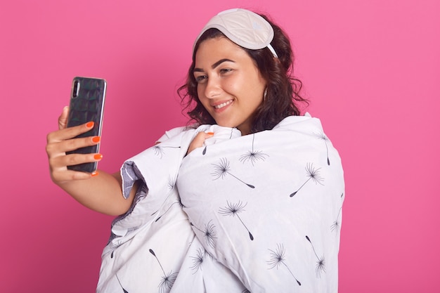 Plan Intérieur De La Belle Jeune Femme Masque De Sommeil Et Couverture Blanche Enveloppée, Prenant Selfie Pendant Le Coucher
