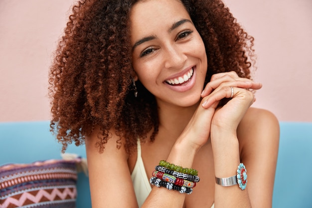 Plan intérieur d'une belle jeune femme métisse à la peau sombre avec une coiffure afro a un sourire agréable, pose sur un canapé, porte un bracelet, entend une histoire drôle d'un ami