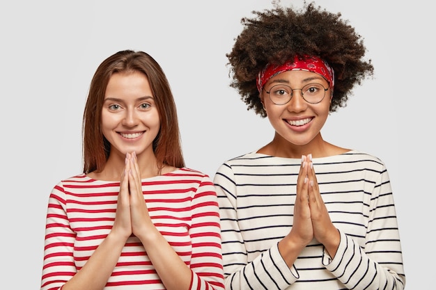 Photo gratuite plan intérieur de belles étudiantes interraciales gaies gardent les mains en signe de prière, demandent bonne chance avant l'examen, portent des chandails à rayures décontractées, se tiennent côte à côte isolées sur un mur blanc