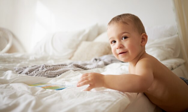 Plan intérieur d'un charmant petit enfant européen d'un an aux joues potelées debout au bord du lit, essayant de grimper, tendant la main pour prendre un livre. Concept de l'heure du coucher, du sommeil et de la garde d'enfants